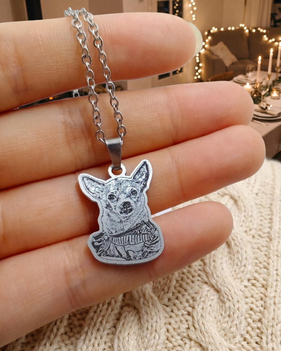 Engraved chihuahua photo necklace held in a hand over a cream sweater with holiday lights behind